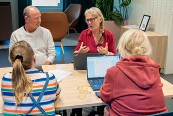 En grupp på fyra personer sitter runt ett bord i samtal, med öppna laptops. En person talar och gestikulerar med händerna.