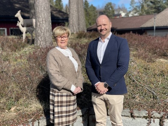 Två personer står utomhus framför byggnader och träd och poserar för en bild.