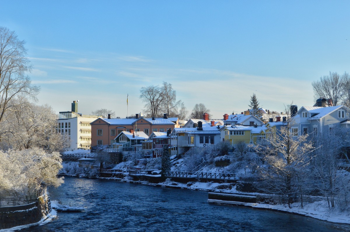 Vintervy över en å i Gävle med snötäckta träd och färgglada trähus längs strandkanten. Himlen är klar och blå, och solen lyser över byggnaderna och det strömmande vattnet.