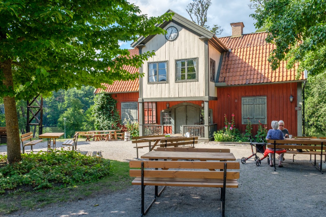 Red wooden house with a tiled roof and a small clock on the gable, surrounded by trees and greenery. In front of the house are wooden outdoor tables and benches on a gravel courtyard. Two older adults sit at a table to the right, one with a walker beside them.