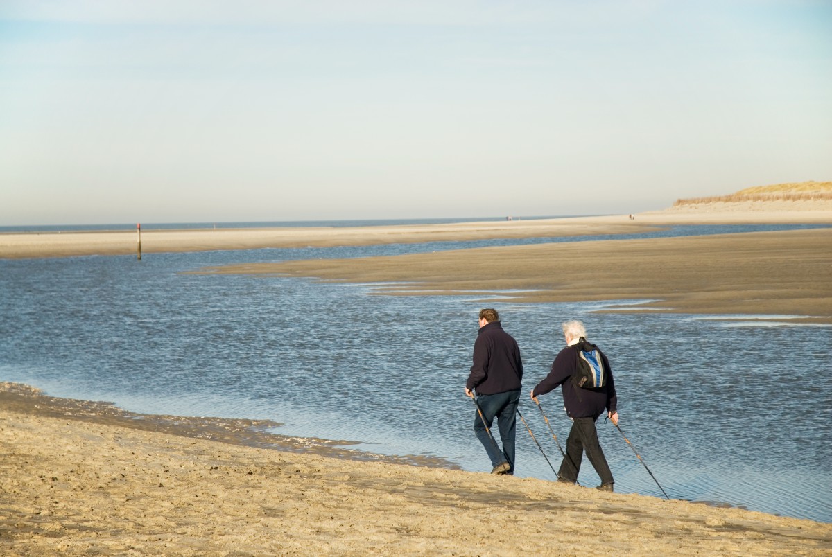 Two older adults walking along a sandy beach beside the water on a clear day.