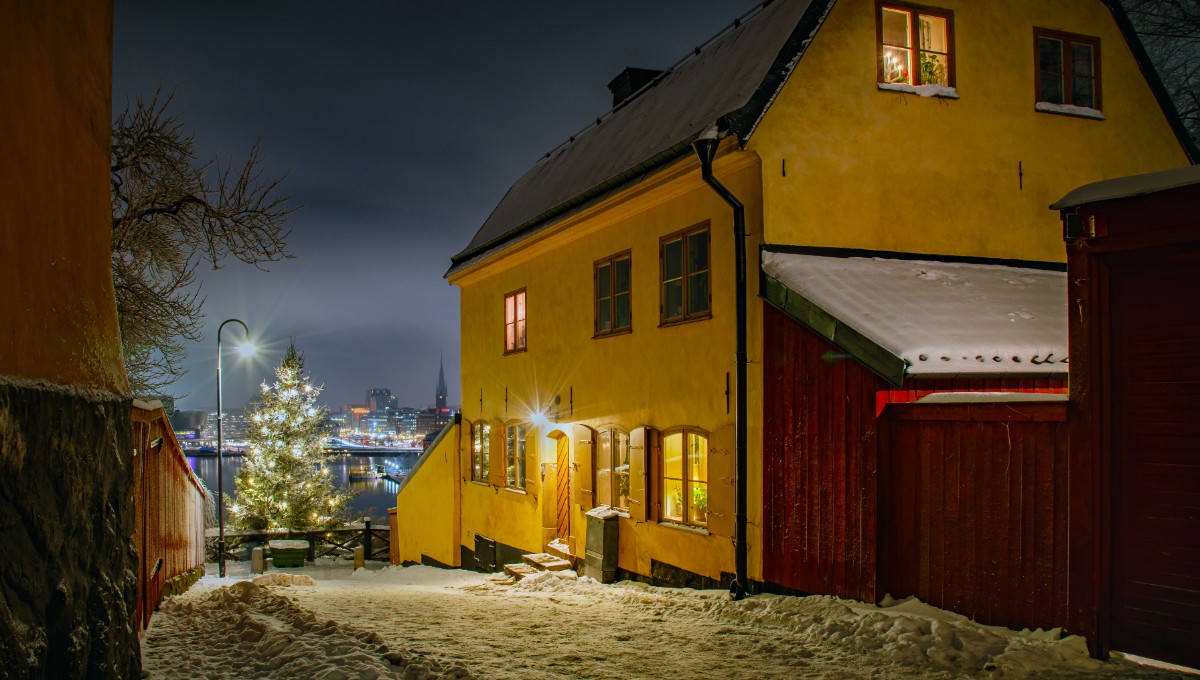En snöig kvällsgata i Stockholm med ett gult hus som lyser inifrån. En pyntad julgran står nära vattnet i bakgrunden.