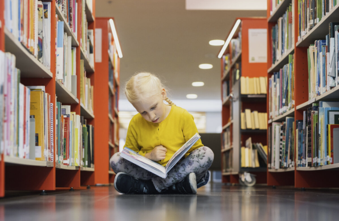En ung flicka sitter på ett biblioteksgolv och läser en bok