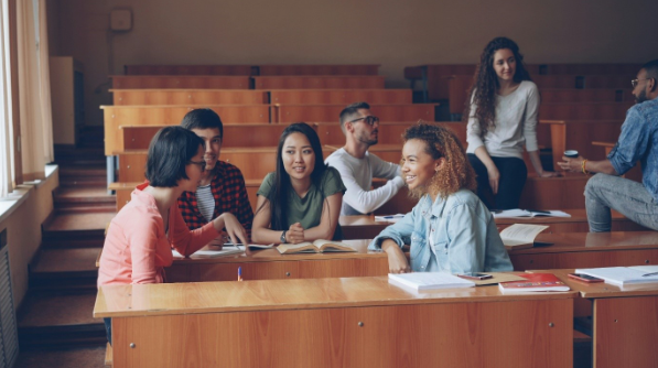 Gruppe af studerende sidder i et auditorium med træborde og taler sammen. Fire studerende sidder samlet foran med bøger og noter på bordet, mens to andre sidder bagved og samtaler. Lyset kommer ind fra vinduerne i siden.