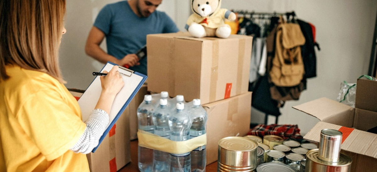 En kvinna med anteckningsblock gör en lista medan en man packar flyttkartonger i ett rum. På bordet står kartonger, flaskor med vatten och konserver, och ovanpå en låda sitter en beige nallebjörn.